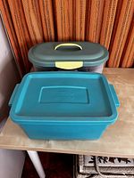 Two closed plastic storage containers, one teal and one translucent with a dark lid and yellow latch, containing sewing threads inside.