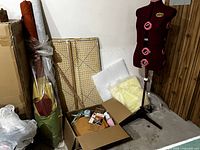 Wide shot showing the mannequin on stand, rolls of fabric standing against the wall, a folding peg board leaning on the wall, and a box on the floor containing sewing accessories and fabric remnants.