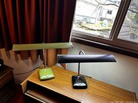 Two vintage desk lamps, one green and one black, side by side on a wood desk near a window, showing overall shape and color.