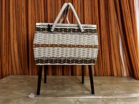 Mid century modern woven sewing basket mounted on four dark tapered wooden legs against an orange striped curtain background.