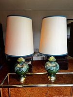 Pair of vintage mid century modern table lamps with ceramic bodies and tall off-white drum shades with blue trim, shown on a glass and brass table.