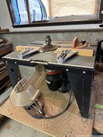 Photo showing the full router table with attached wooden measuring jigs and an orange guide accessory on top. The table surface is ribbed metal. The table legs are black metal with a sturdy build.