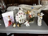 Full lot photo showing all figurines arranged on table: Willow Tree figurine holding lilies, Royal Doulton Kathryn figurine in floral dress, fairy with butterfly, cherub angel, and two small bird figurines.