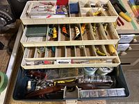 Top view of open tackle box showing fishing lures organized by compartments on the top tiers, along with lures and small accessories in the lower section of the box.
