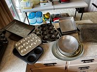 Multiple muffin tins, round and square cake pans, rectangular trays, and wire cooling racks arranged on a kitchen counter.