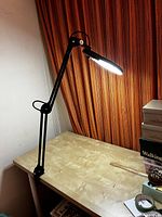 Rotatable LED magnifying lamp lit up on desk with orange-patterned curtain in the background.
