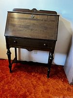 Front view of wooden secretary desk showing hinged drop-front writing surface closed, decorative backboard, and drawer below desk surface.