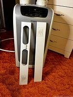 Front view of the Simplicity portable AC unit showing its silver and gray housing, front air vents, and removable panels leaning beside it.