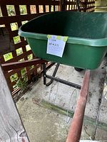Green plastic True Temper wheelbarrow with wide tray and attached metal frame with wheel under, taped lot label visible on the tray front.
