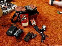 Lot showing all items including Canon Sure Shot camera with case, Nikon CoolPix camera, Kodak Trimlite Instamatic camera in packaging, Stitz tripod, battery charger, cables, and brown carrying bag.