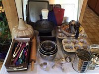 Full lot setup showing an assortment of metal bakeware pans, cooling racks, flour sifters, utensils, and rolling pin
