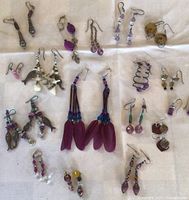 Overview of assorted beaded earrings laid out on a light cloth, displaying variety of colors mostly purples, pinks, and browns, highlighting the feathered pair in the center