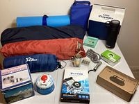 Wide view of camping gear laid out including rolled tents, foam sleeping pad, cooler, thermos, cookware in box, and assorted camping accessories.