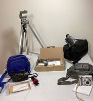 Photo showing the Canon camcorder on black bag, Canon Powershot camera on gray bag, Yashica camera in blue bag, small tripod and box with camera accessories like charger, cables, manuals, and remote control