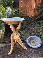 Side view of the birdbaths on ground showing the ceramic birdbath with tripod stand and round metal hanging birdbath lying next to it.