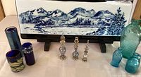 Full arrangement with blue and white mountain scene glass panel on wooden stand, several small blue glass vases, three tiny ornate glass bottles, and mini blue jug vase.