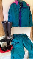 Photo of teal North Face Gore-Tex ski jacket hanging on wall, teal ski pants on table, black ski gloves, red framed ski goggles, grey boots, and grey rabbit fur ear muffs on white surface