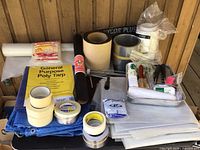 Overview of tarps, plastic sheeting, masking tapes, paint trays, and other painting supplies on wooden surface.
