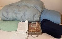 Two blue down blankets on bed with plush navy throw blanket, white and light green sheets, and safari-themed sheet set in packaging