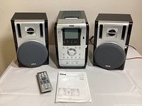 Full view of RCA portable stereo system including central unit and two speakers, with remote control and user manual placed in front