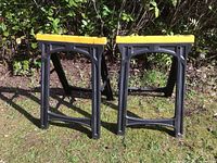 Two folding plastic sawhorses with black legs and yellow tops standing upright on grass against a bushy background.