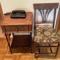 Front view of the small wooden cabinet with woven rattan top holding the black vintage phone, and wooden chair with floral upholstered seat beside it.