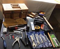 Wooden toolbox with metal latches, leather gloves, screwdrivers, wrench set, hammer, and other hand tools on a table.