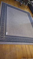 View of entire rectangular grey area rug with a blue Greek Key border on a wood floor.