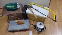 Photo showing all items on floor: yellow Coleman cooler with axe and saw on top, grey stove cases, pots, folding stool, black radio light, and sleeping mat bag.