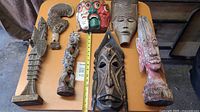 Seven carved wooden masks and figures arranged on a table, showing variety in size, style, and color.