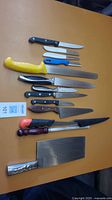 Photo showing various knives on a table including cleavers, fillet knife, serrated knives, and sharpening rod all arranged for display.