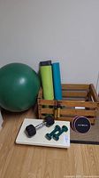 Wooden crate filled with three rolled yoga mats, peach fitness bands sliders, vintage skipping rope with wooden handles. Large green exercise ball adjacent to crate. Two 3 pound green dumbbells and one 15 pound black dumbbell set on white step-up board on floor.