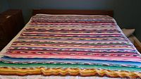 Crocheted rainbow afghan blanket fully spread out on a bed, showing striped multicolor pattern and scalloped edge