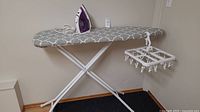 Ironing board with iron and hanging dryer positioned in corner, showing full ironing board with patterned cover and hanging dryer clipped to side.