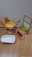 Yellow baby doll pram, smaller floral fabric stroller, and white plastic bathtub shown together on wood floor.