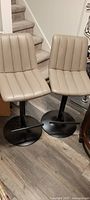 Two beige adjustable bar stools side by side, showing vertical tufting and lever mechanisms.