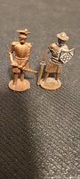 Two small copper soldier figurines standing upright on a black textured surface. The soldier on the left has a distinct hat and sword, the soldier on the right has a shield with a diamond pattern and a hat.