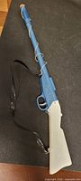 Full view of the vintage cork toy rifle with blue barrel and white stock, showing length and attached strap.
