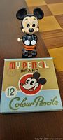 Front view of Mickey Mouse wind-up toy standing behind the box of My Pencil Brand colour pencils showing both items clearly.