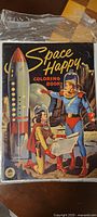 Front cover of the Space Happy coloring book showing two space cadet characters in futuristic suits and helmets with a rocket ship in the background.