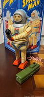 Front view of the tin space man astronaut toy standing with the original box in the background, remote control on table.