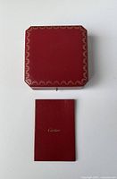 Top view of closed red Cartier jewelry case box with gold decorative border. Includes small red Cartier booklet below.