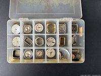 Overall view of plastic compartment box holding 18 separate vintage watch movements, dials, and cases in various shapes and sizes, showing watch components closely packed.