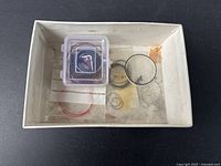 Top view of white cardboard box with assorted vintage men's watch gaskets including thin and thick round bands, red, black, and grey colors, and a small transparent plastic container holding smaller parts inside the box.