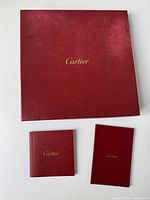 Top view of closed square red Cartier box with gold logo in center, accompanied by two small red booklets with Cartier gold text