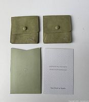 Two green suede jewelry pouch holders with snap button closure, authenticity certificate and branded information booklet placed on white background.