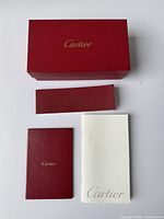 Red Cartier sunglasses box with gold logo on lid and three Cartier booklets in red and white.