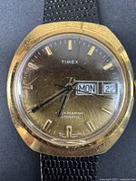 Full front view of gold-tone Timex men's watch showing dial, hands, day and date window, and black strap