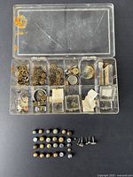 Clear plastic organizer with multiple compartments containing various small metal watch parts including gears and screws. Below the organizer are about 30 assorted vintage pocket watch crowns laid out in rows.