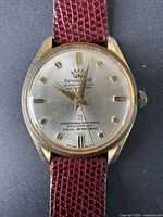 Front view of vintage Remington watch showing gold-tone case, white dial, gold markers and hands, and red leather strap.
